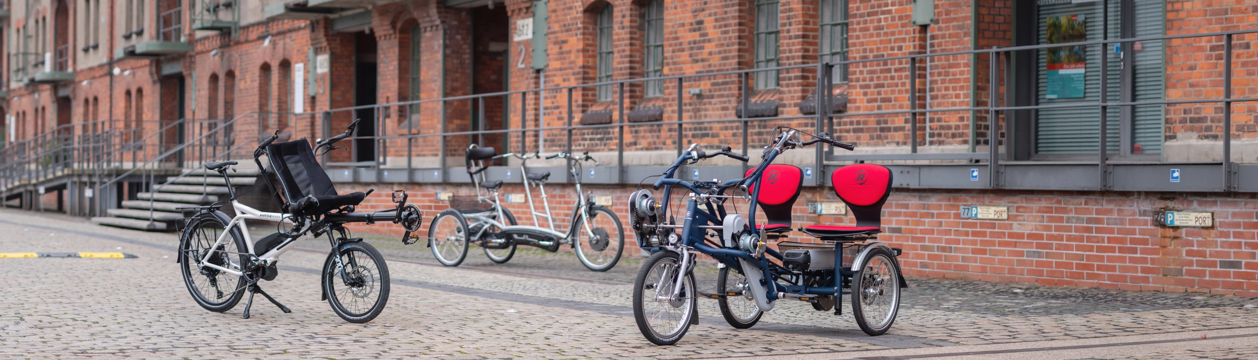 Ein Paralleltandem, Tandem mit Liegefunktion und Dreiradtandem stehen vor einem Gebäude. Könnte ein alter Hafen sein. 