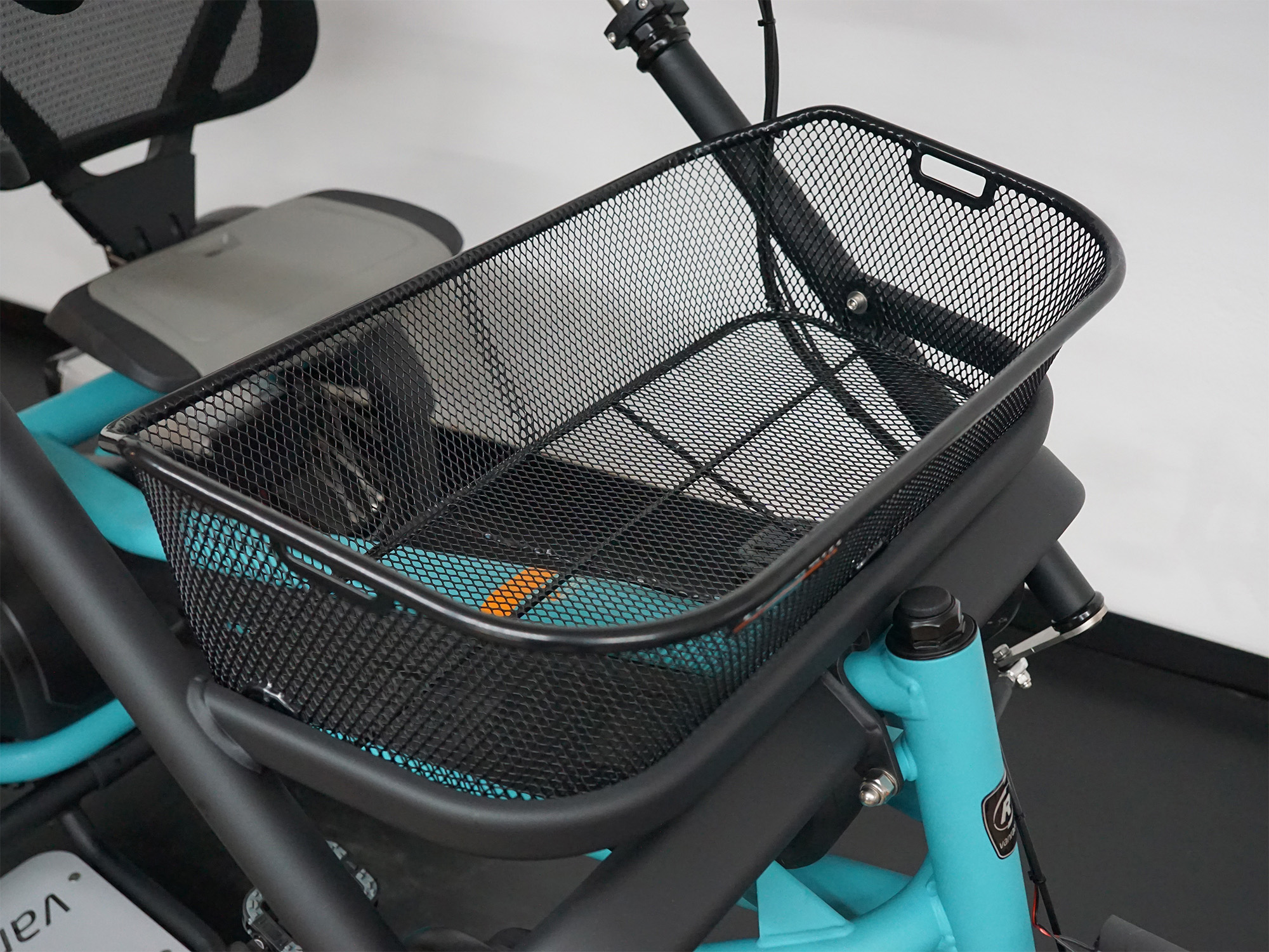 Dreirad mit schwarzem Metallkorb vorne und türkisfarbenem Rahmen, Sitz mit Rückenlehne im Hintergrund.
