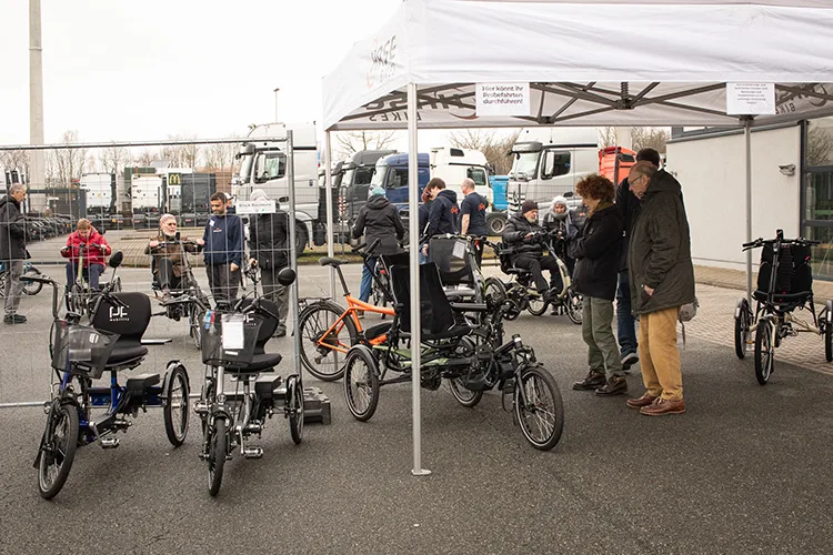 Mehrere Spezialfahrräder und Dreiräder sind unter einem weißen Pavillon aufgebaut. Menschen stehen daneben, probieren die Räder aus oder schauen sie sich an.