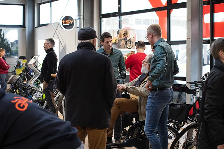 Ein älterer Mann sitzt auf einem Spezialfahrrad, während zwei Berater mit ihm sprechen. Andere Besucher schauen sich im Hintergrund Fahrräder an.