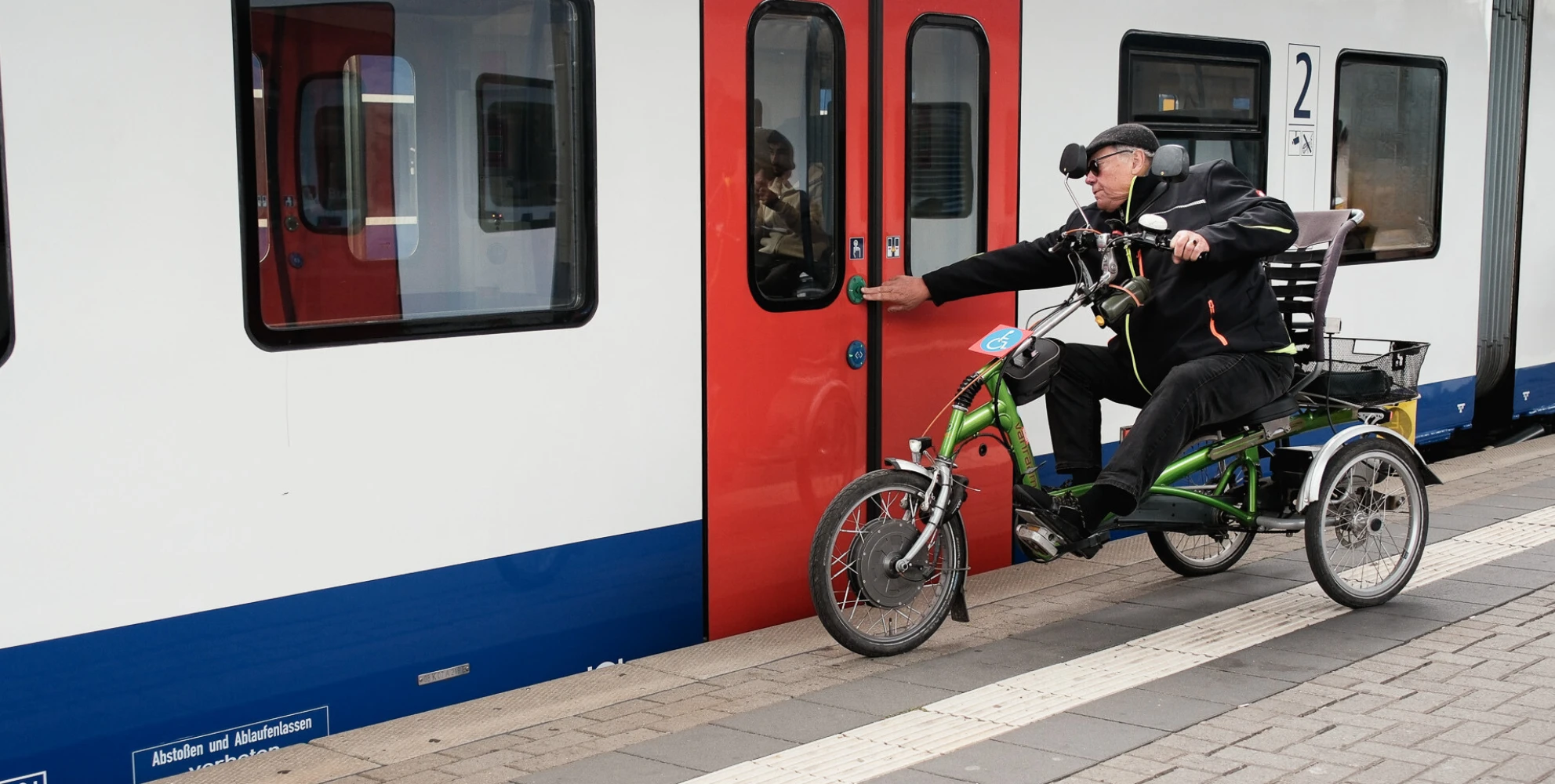 Mann im Dreirad öffnet eigenständig die Tür eines Regionalzugs am Bahnsteig.