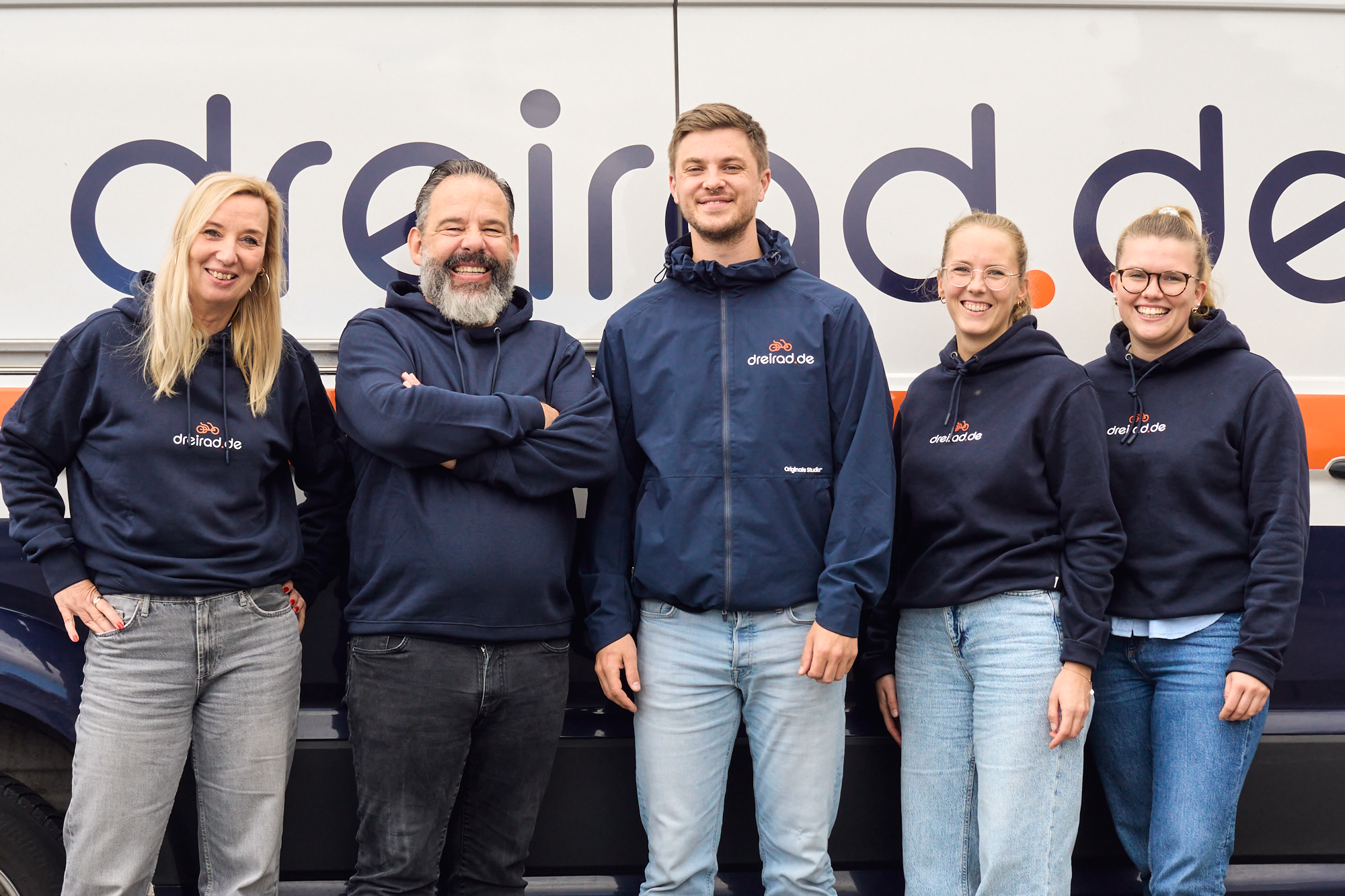 Drei Frauen und zwei Männer stehen lächelnd vor einem Fahrzeug mit dem dreirad.de Logo und lächeln