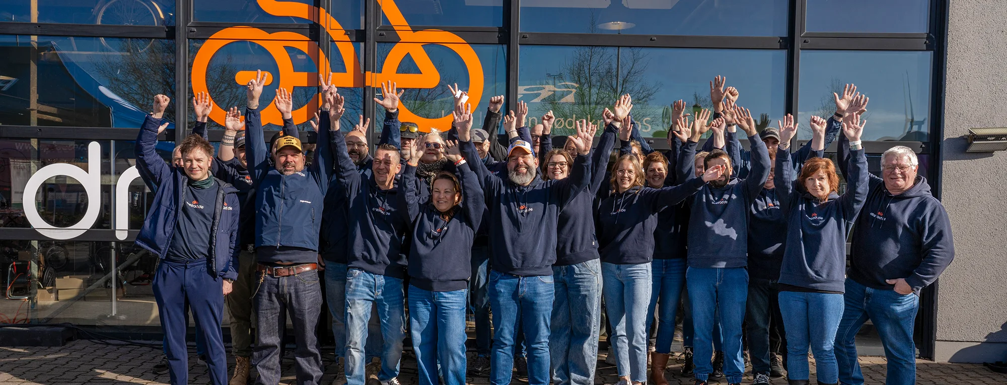 Team von dreirad.de steht gemeinsam vor einem Geschäft und winkt in die Kamera.