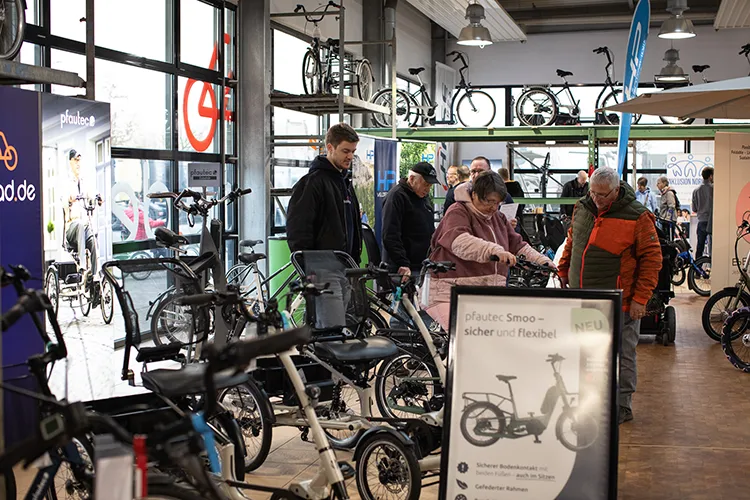 Menschen in einer hellen Halle betrachten E-Bikes und Spezialfahrräder. Ein Mann erklärt etwas, während andere Besucher verschiedene Modelle anschauen.