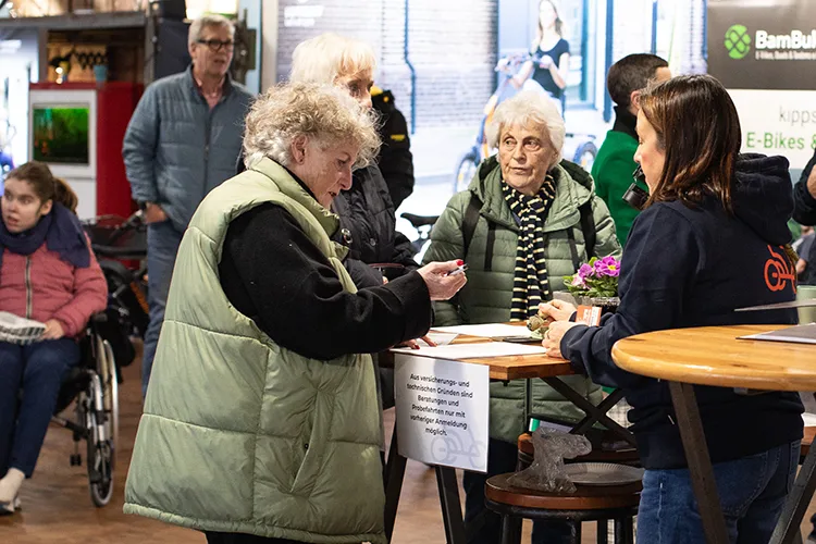 Zwei ältere Frauen stehen an einem Holztisch und schreiben etwas auf, während eine Mitarbeiterin ihnen hilft. Im Hintergrund sitzen weitere Besucher und unterhalten sich.