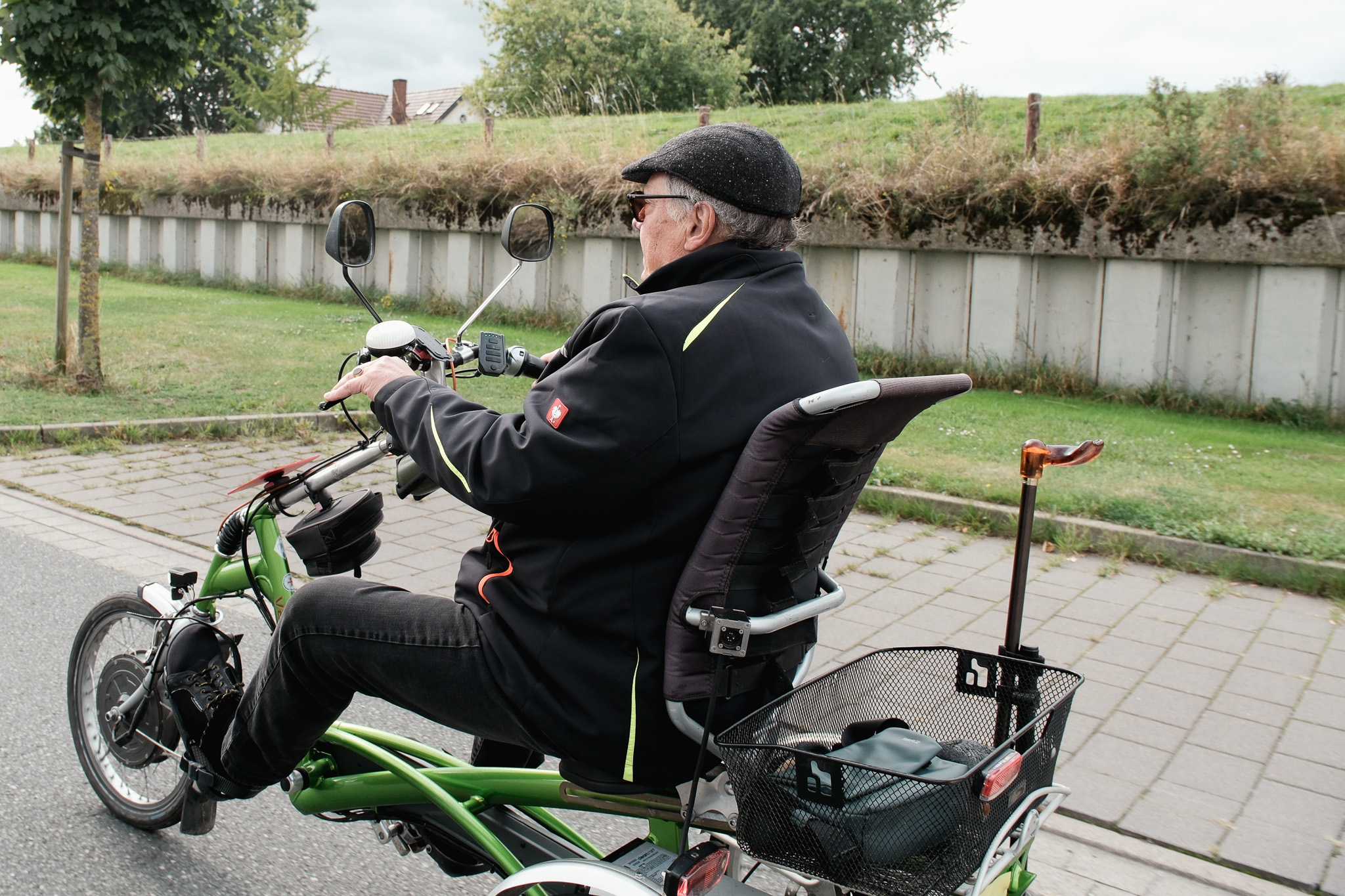 Älterer Mann fährt auf Reha-Dreirad mit hoher Lehne, Spiegeln und Gehstockhalter.