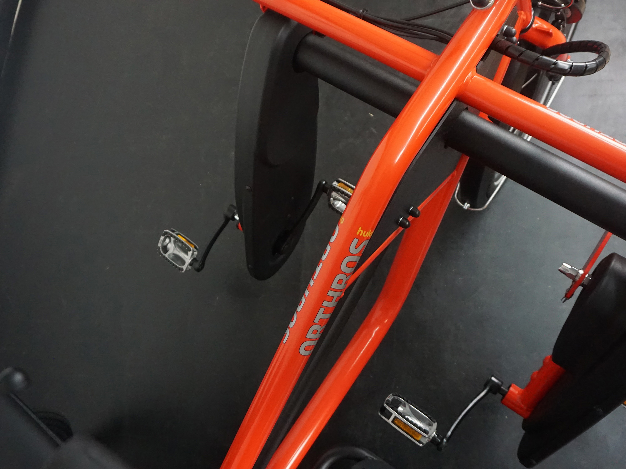 Oberansicht eines orangenen Orthros-Tandems mit zwei Tretlagern und nebeneinanderliegenden Pedalen.